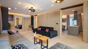 A modern living room with a sectional sofa, two wooden side tables, a black sofa with pink cushions, and a kitchen area with a countertop and chairs. - Hedera Estate, Villa Hedera XVII (Dubrovnik)