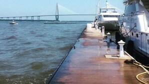 A waterfront dock with yachts moored alongside, a bridge in the background, and calm water.