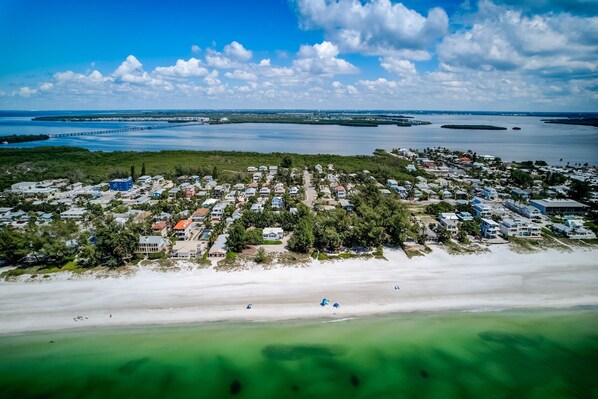 An aerial view of a coastal residential area with houses, a beach, and a bridge in the background.