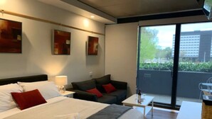 A hotel room with a large bed, a sofa, a small table, and a balcony with a view of the city.
