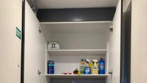 A compact laundry room with a front-loading washing machine, shelves with cleaning supplies, and a red bucket.