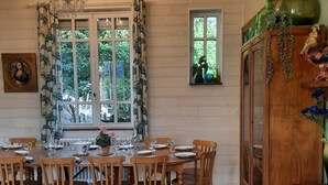 A dining area with a wooden table set for six, surrounded by chairs, a chandelier, and a window with curtains. - La Villa Luchonnaise, 5mn du Centre Ville, Télécabine et Thermes (Bagnères-de-Luchon)