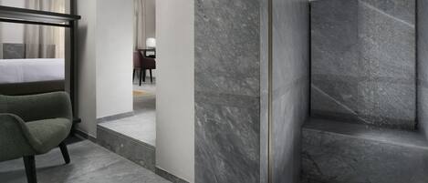 Habitación de hotel moderna con una cama grande, un sillón verde, pared de ducha de mármol y un espejo amplio.