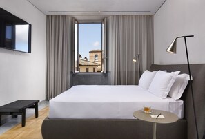 Una habitación de hotel moderna con una cama grande, una mesita con una taza y un bolígrafo, un televisor montado en la pared y una ventana con cortinas.
