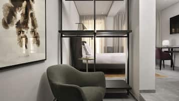 A modern hotel room with a green armchair, a bed with white linens, a desk with a lamp, and a large framed artwork on the wall.