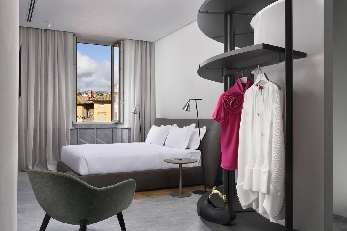 A modern hotel room with a bed, a small round table, a chair, a wardrobe, and a view of buildings outside.