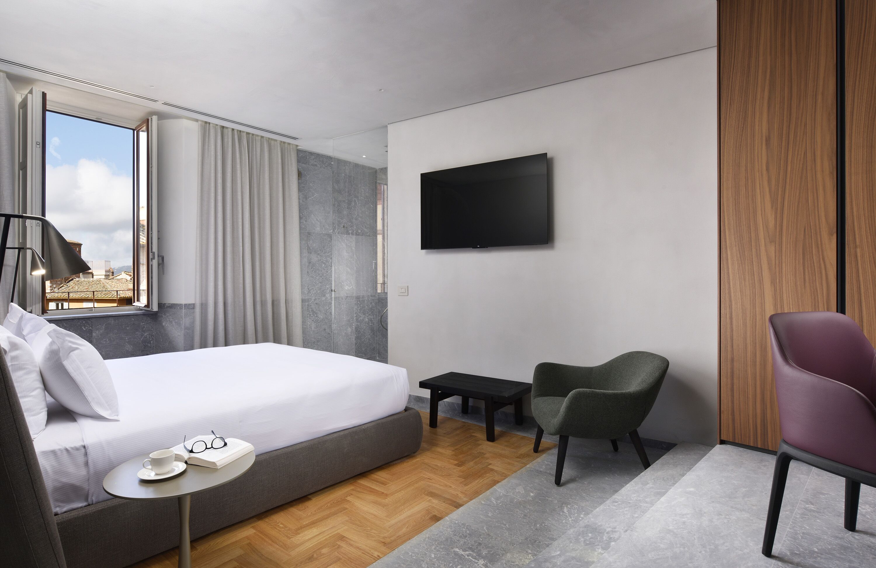 A modern hotel room with a large bed, a flat-screen TV, a small table, two chairs, and a view of the city through a large window.