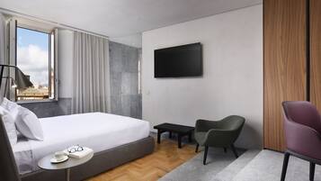 Una habitación de hotel moderna con una cama grande, un televisor de pantalla plana, una mesita pequeña, dos sillas y vistas a la ciudad a través de un ventanal amplio.