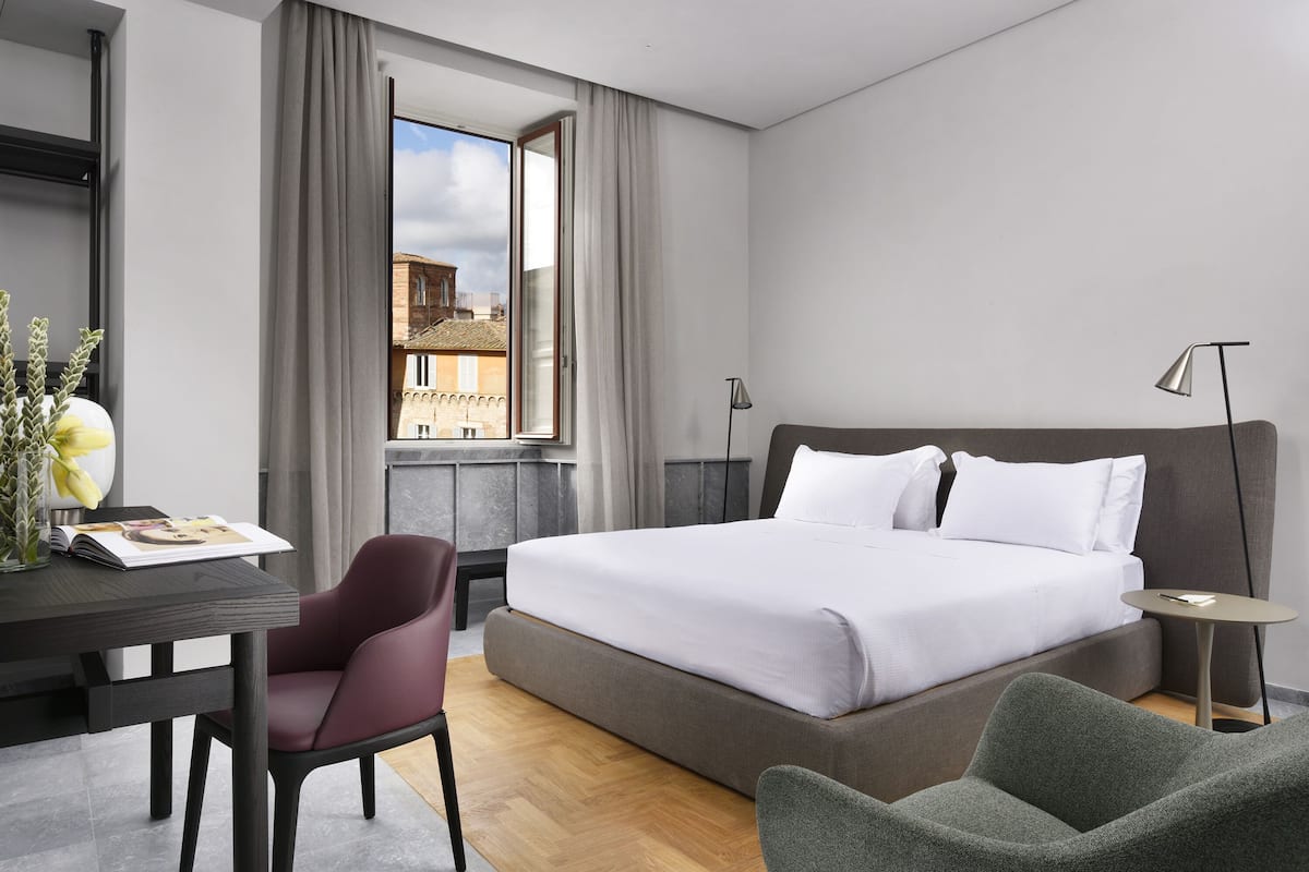 A modern hotel room with a large bed, a desk with a chair, a small table, and a view of the city.