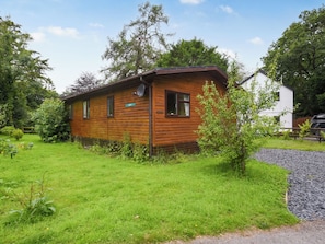 A wooden cabin with a small porch, surrounded by a grassy yard and trees.