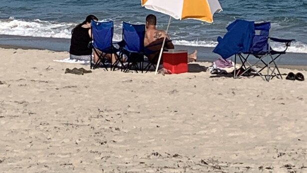 A beach scene with a person under an umbrella, two blue beach chairs, and a cooler on the sand.