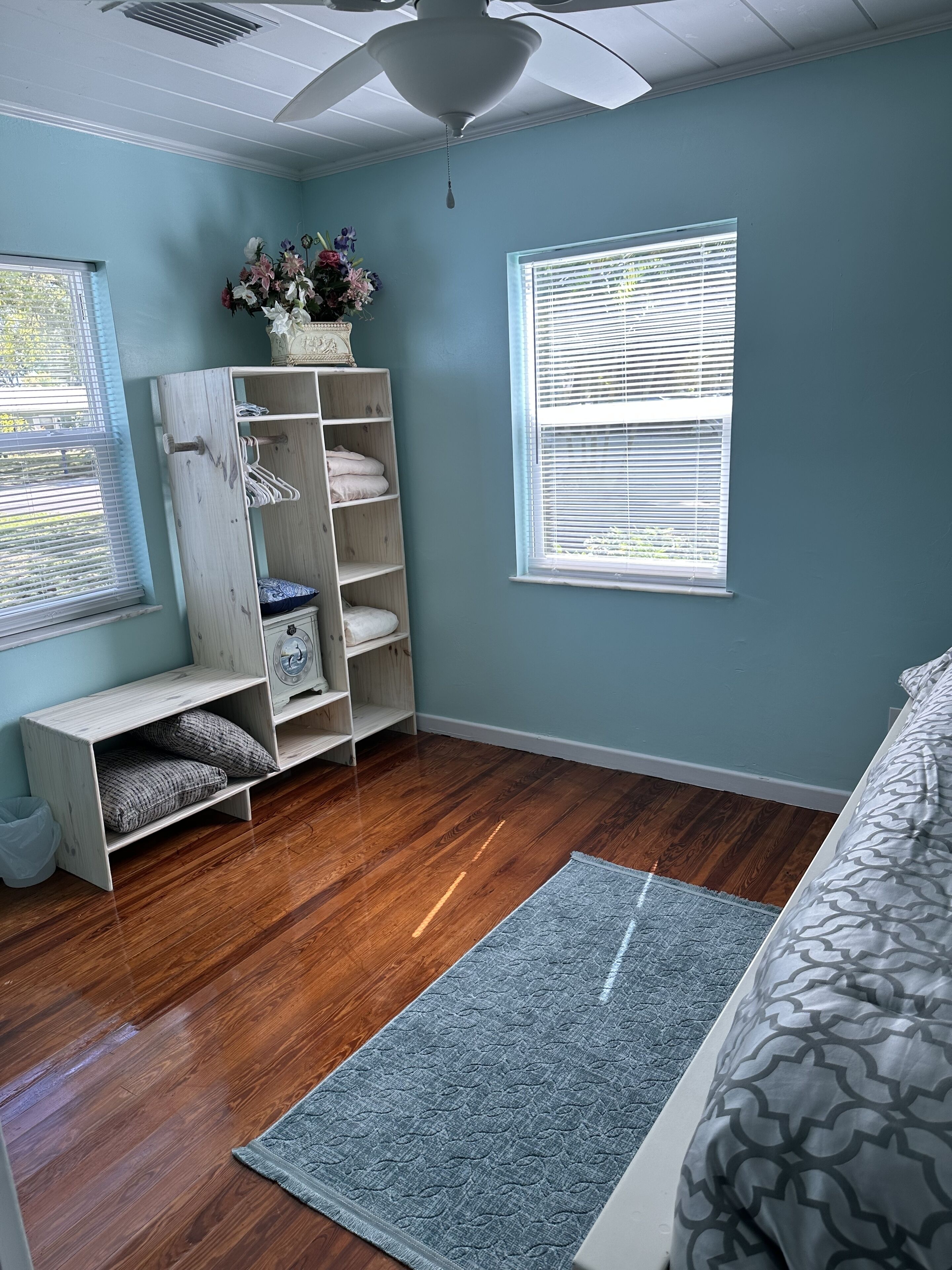 Une chambre avec un plancher en bois, un lit avec une literie à motifs, un tapis gris, une étagère blanche avec des vêtements pliés et une fenêtre avec des stores.