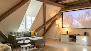Un salon chaleureux de style loft, doté d’un haut plafond, de poutres apparentes, d’un grand téléviseur à écran plat et d’un coin salon confortable avec un canapé et un pouf.