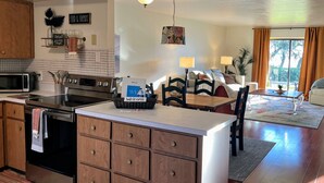 A modern kitchen with a central island, wooden cabinets, and a microwave. There is a dining area with a table and chairs, and a view of the outdoors through large windows.