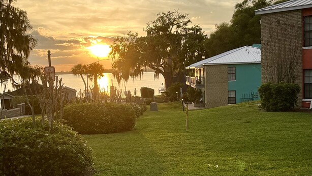 A sunset view over a body of water with a 'No Parking' sign, a grassy area, and buildings with brick and metal roofs.