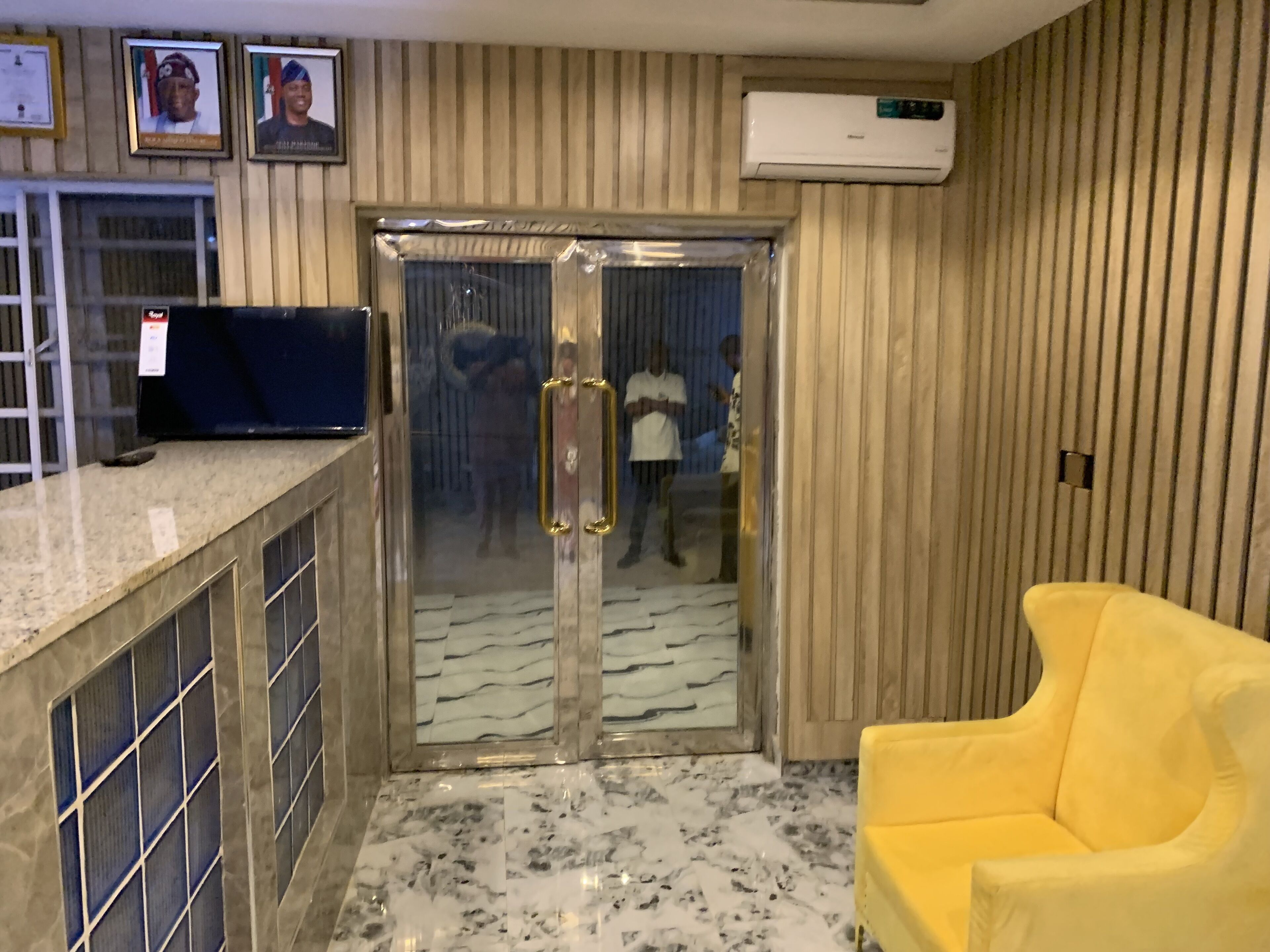 Zona de recepción con piso de mármol, un sillón amarillo, mostrador de recepción, certificados enmarcados y un equipo de aire acondicionado.