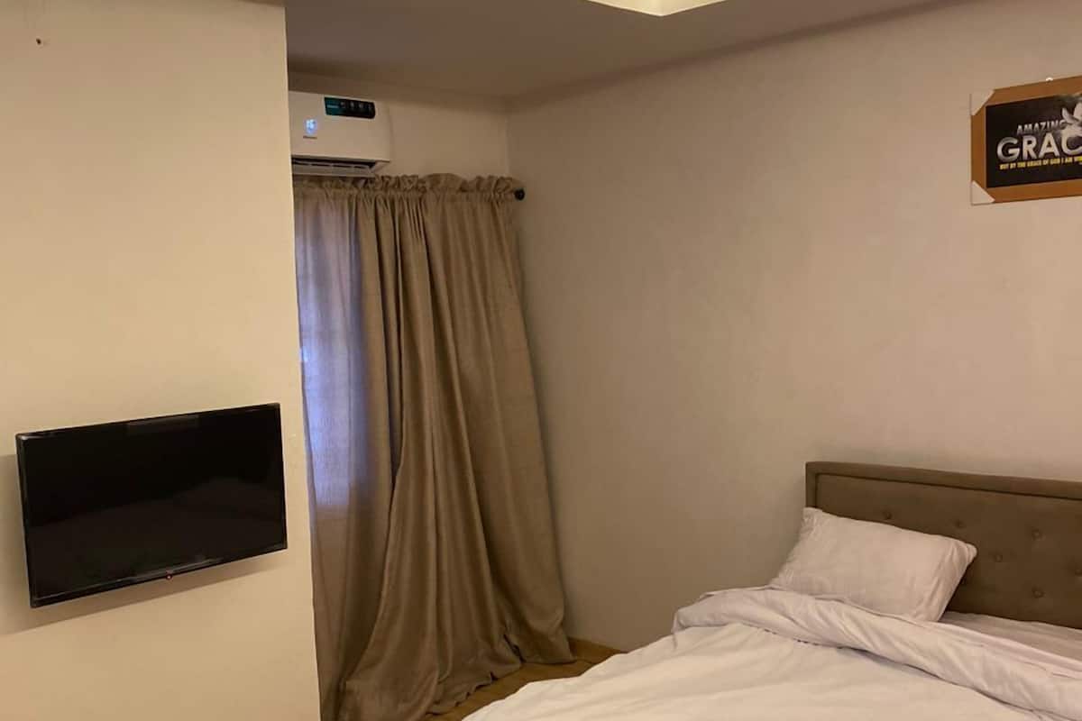 A hotel room with a bed, a flat-screen TV mounted on the wall, a ceiling light, and a window with curtains.