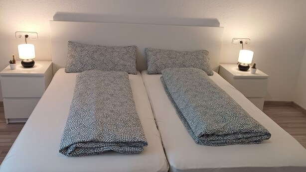 A double bed with two nightstands, each with a lamp, in a room with a plain white wall and wooden flooring. - Gemütliche Ferienwohnung mit Kamin (Emmingen-Liptingen)