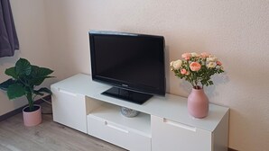 A modern living room with a flat-screen TV on a white cabinet, a potted plant, a vase with flowers, and a wall clock. - Gemütliche Ferienwohnung mit Kamin (Emmingen-Liptingen)