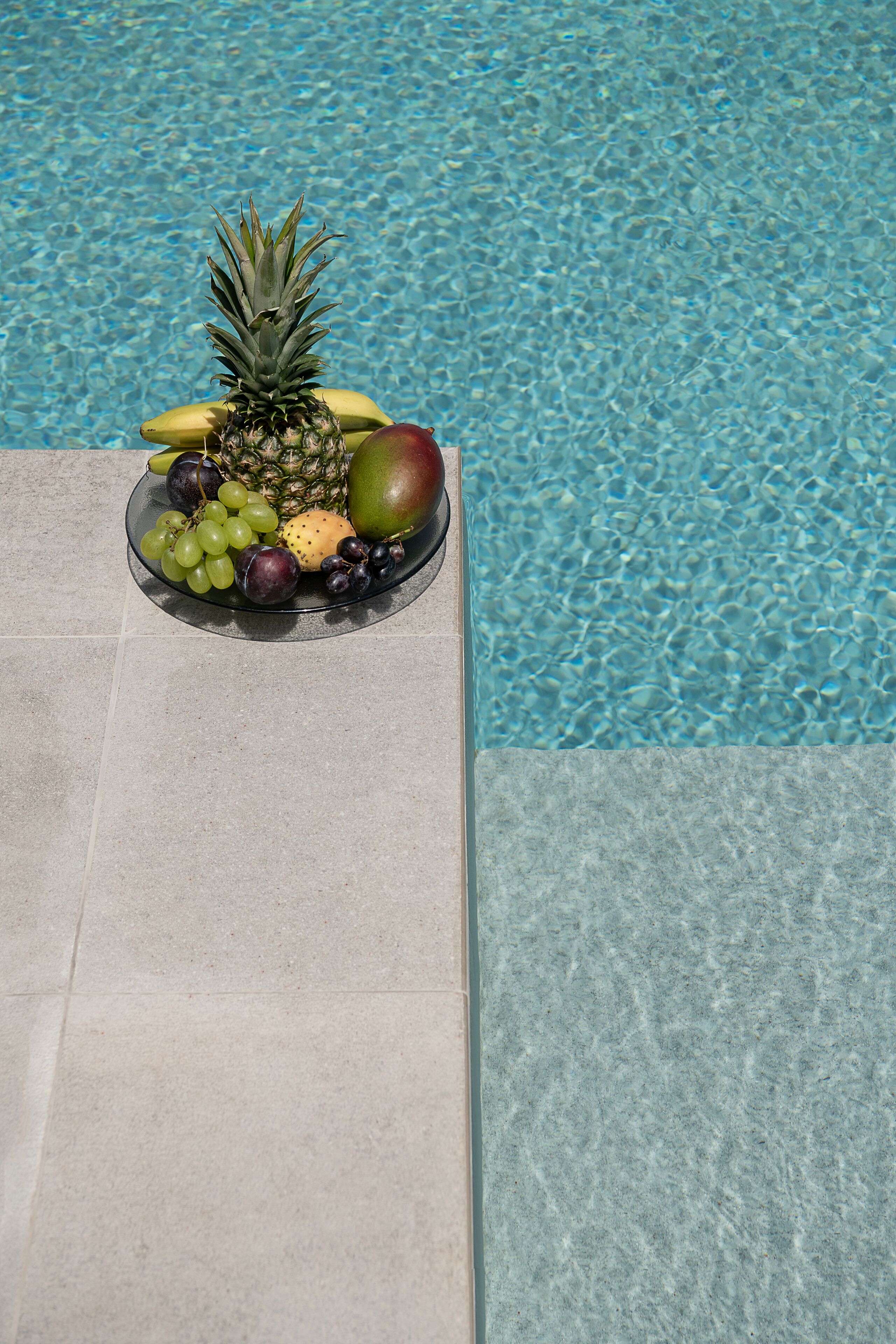 A fruit bowl with a pineapple, banana, mango, grapes, and plums by a pool.