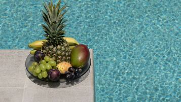 A fruit bowl with a pineapple, banana, mango, grapes, and plums by a pool.
