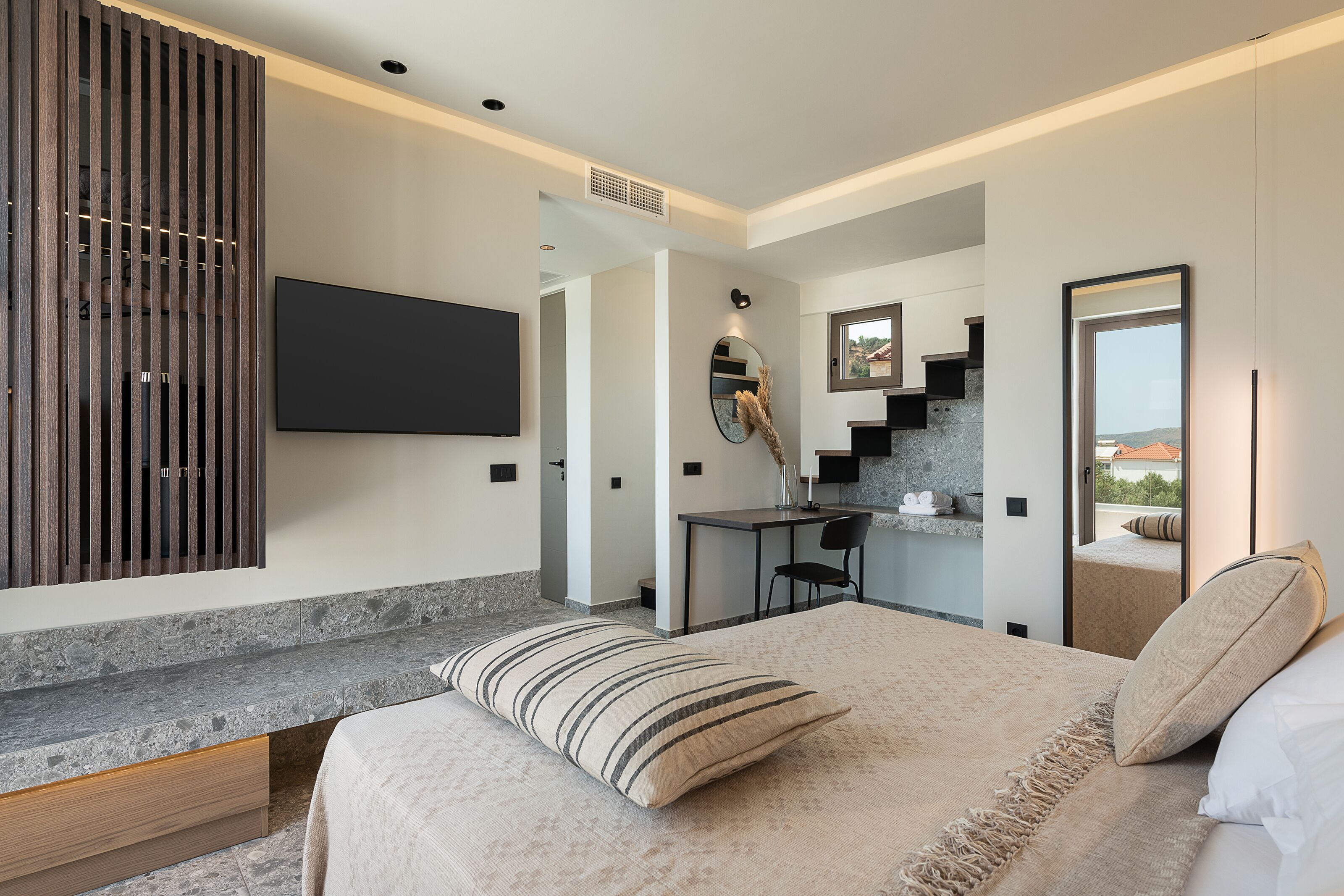 Ein modernes Schlafzimmer mit einem großen Bett, einem an der Wand montierten Fernseher, einem Schreibtisch und Blick ins Freie.