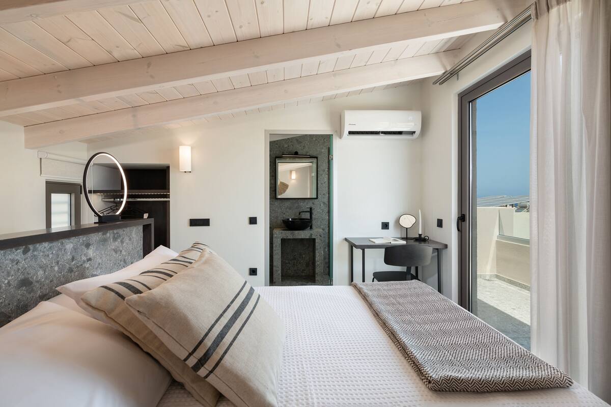 Habitación de hotel moderna con cama, escritorio pequeño, espejo y vista de la ciudad a través de un ventanal amplio.