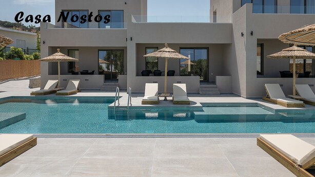 A modern pool area with lounge chairs and umbrellas, a clear sky, and the text "Casa Nostos". - Casa Nostos (Chania)