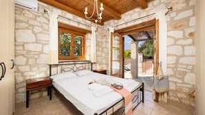 Une chambre avec un mur en pierre, un plafond en bois, un lit avec du linge de lit blanc, une table de chevet et une fenêtre avec des rideaux.