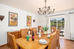 Comedor con mesa de madera dispuesta para seis personas, rodeada de sillas de mimbre. Hay dos carteles en la pared, un candelabro sobre la mesa y vistas al exterior a través de un gran ventanal.