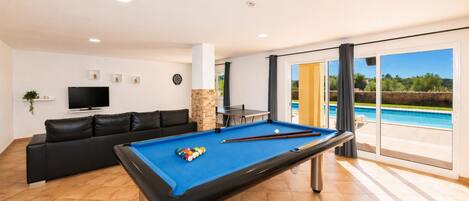 A pool table in a room with a sofa, TV, and a view of a swimming pool.