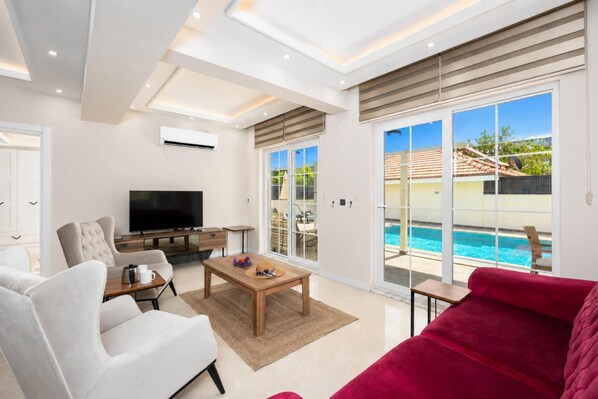 A modern living room with a TV, sofa, coffee table, and a view of a swimming pool.