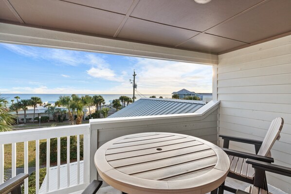 A balcony with a round wooden table and a chair, overlooking a beach with palm trees and a house in the background. - Destin Retreat*Heated Pool*Gulf Views*Beach Gear (Destin)