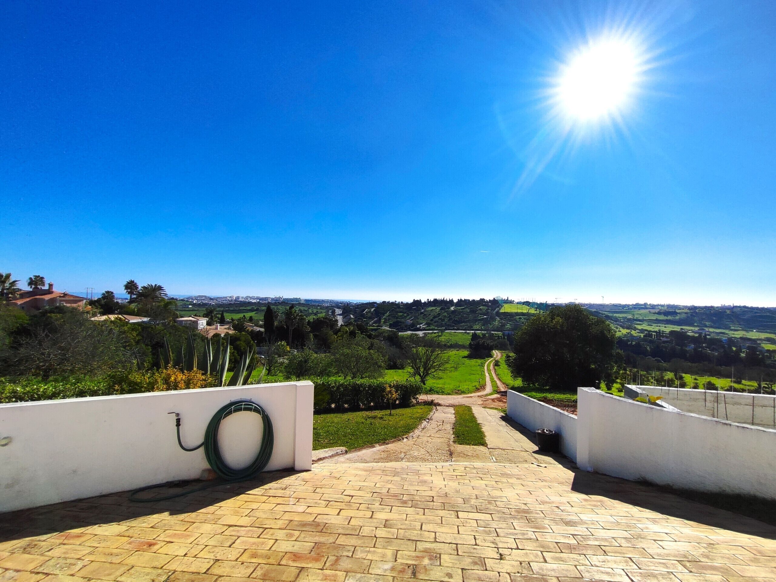 Un camí pavimentat que condueix a una vista panoràmica d'un paisatge verd amb un cel blau clar i un sol brillant.