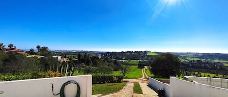 A paved pathway leading to a scenic view of a green landscape with a clear blue sky and a bright sun.
