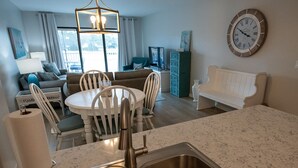 Una cocina moderna con un lavamanos, una mesa de comedor y sillas. Hay una sala de estar con un sofá y una televisión. Un reloj está en la pared.