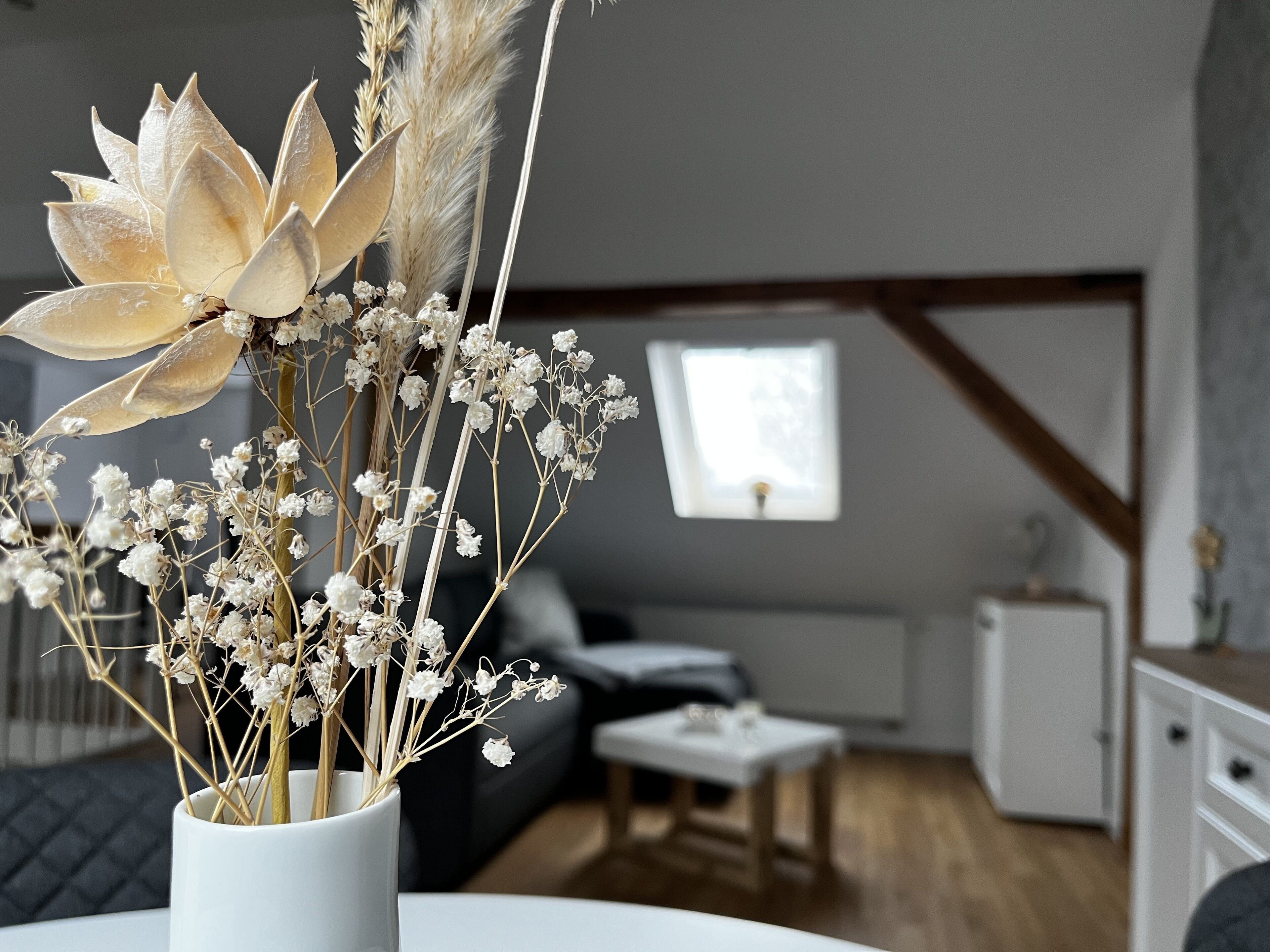 Un salon avec un vase blanc contenant des fleurs séchées et des plumes d'herbe des pampas. On y trouve une fenêtre donnant sur l'extérieur, un canapé blanc, une petite table et un meuble blanc.