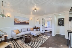 A modern living room with a sectional sofa, a coffee table, and a rug. There is a painting on the wall and a door leading to another room.