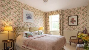 A bedroom with floral wallpaper, a bed with a patterned bedspread, two bedside lamps, a window with curtains, a chair with a vase of flowers, and a framed picture on the wall. - 2 bedroom accommodation in Broughton, near Skipton (Broughton, near Skipton)