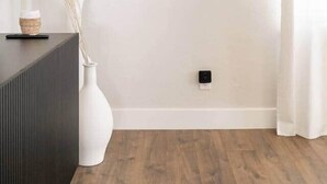 A minimalist room with a framed artwork on the wall, a white vase with dried plants, a dark wooden sideboard, and a white curtain.