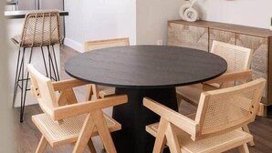 A modern dining area with a round table, four wooden chairs, a white hanging light fixture, a woven wall decoration, and a kitchen area with a refrigerator.