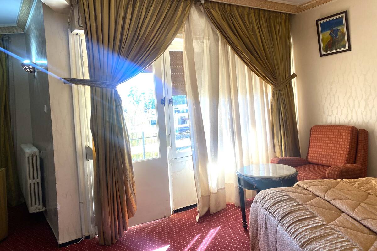 A hotel room with a bed, a chair, a small table, and curtains.