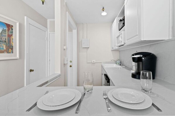 A modern kitchen with white cabinets, a coffee maker, and a dining table set for two. - Pool/StepsToBeach/Dine/Pearl 1brCondo (Palm Beach)