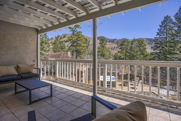 A covered outdoor area with a sofa and a table, a balcony with a railing, and a view of trees and hills. - Stone Statesroom (Cascade)