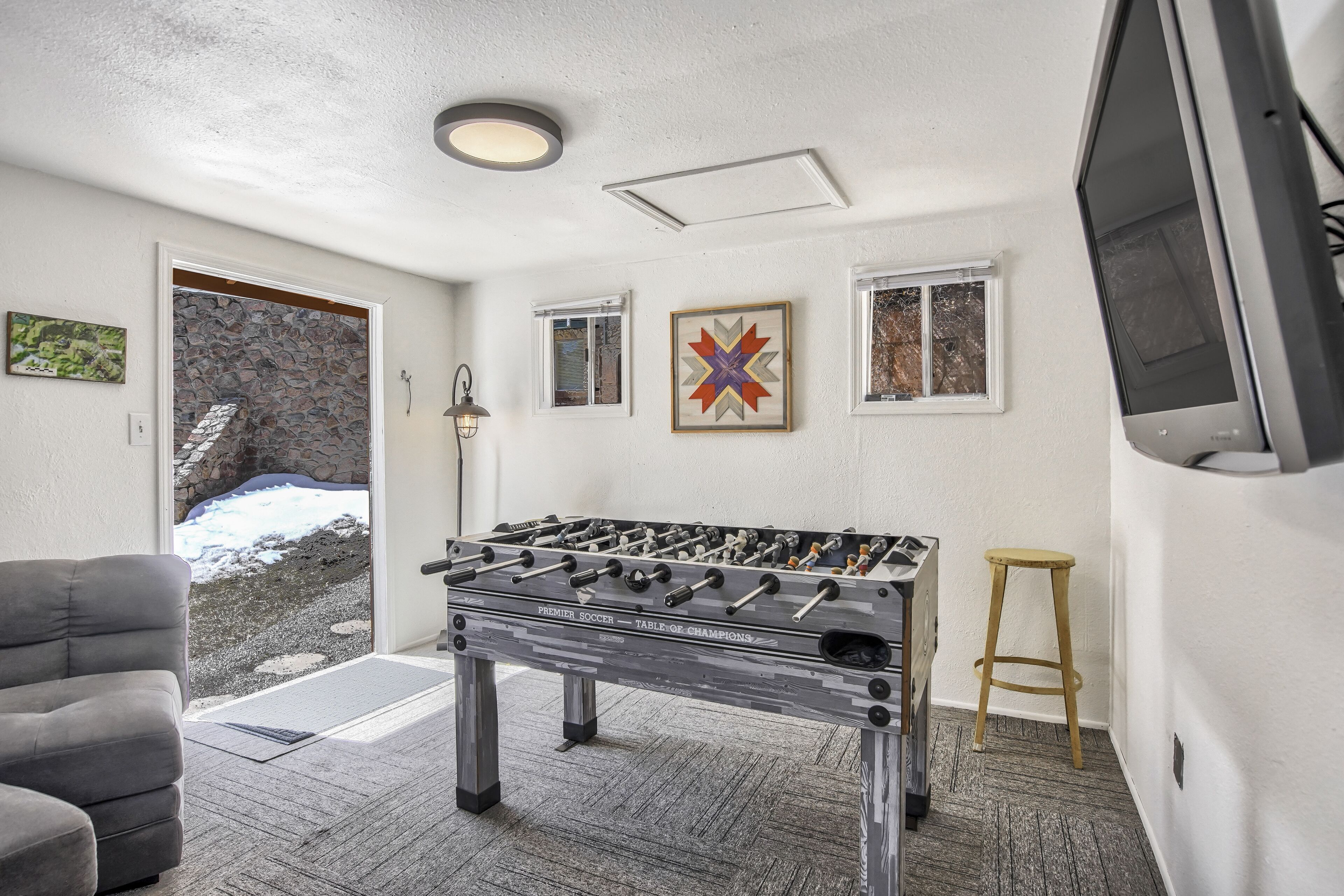 A room with a foosball table, a gray sofa, a wooden stool, and a stone wall with a window.