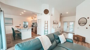 A modern living room with a teal leather sofa, a kitchen in the background, and a dining area with a small island. - Lakefront Naples Lely Resort, 3BR Heated Pool + Players Club & Spa Access Option (Naples)