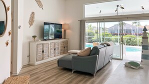 A spacious living room with a large flat-screen TV, a sofa, a wooden cabinet, and a view of a swimming pool through large windows. - Lakefront Naples Lely Resort, 3BR Heated Pool + Players Club & Spa Access Option (Naples)