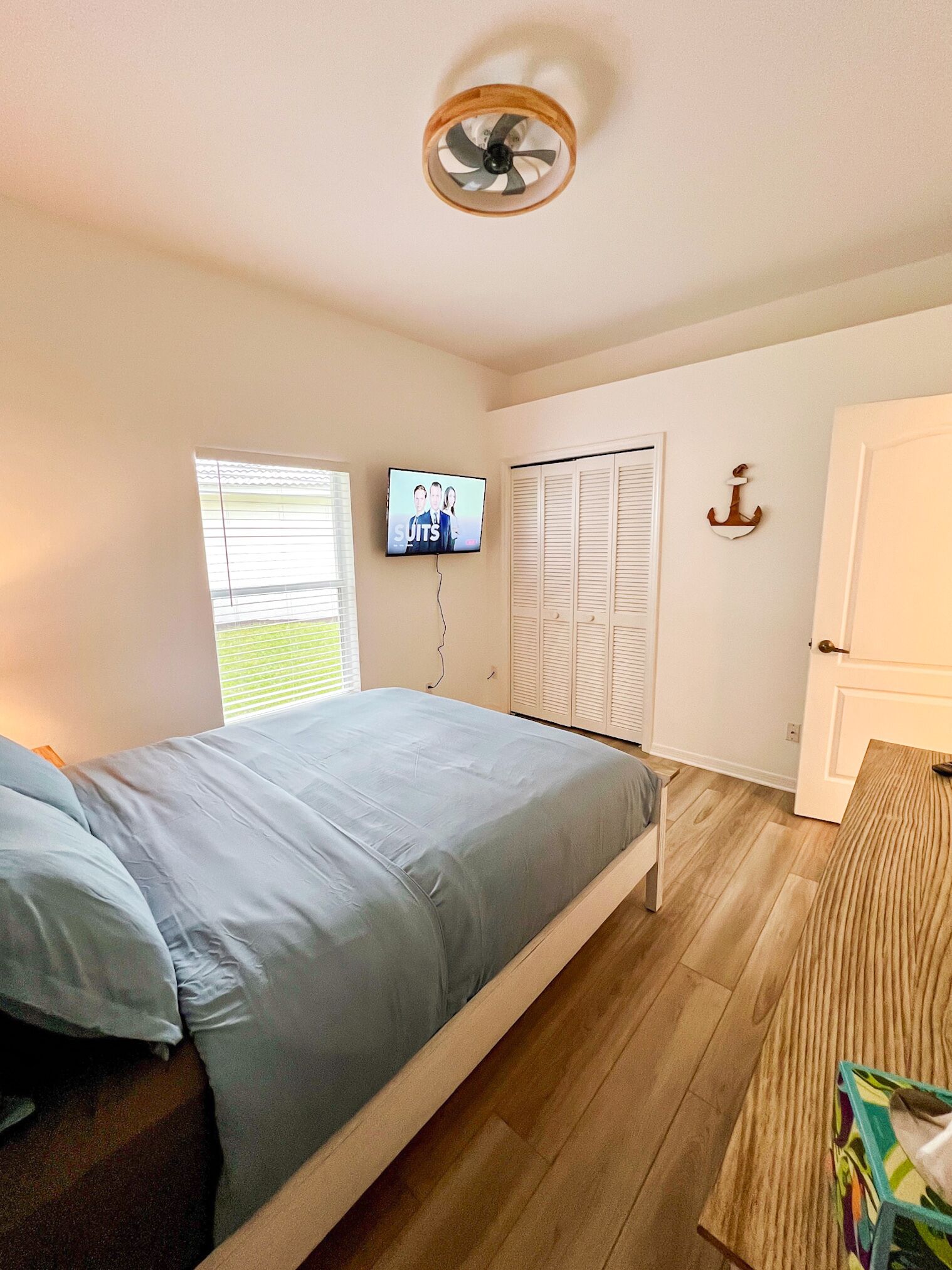 Un dormitorio con cama, televisor montado en la pared, ventana con persianas, puerta y ventilador de techo.