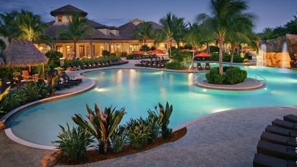 A resort pool with lounge chairs, palm trees, and a building with a thatched roof. - Lakefront Naples Lely Resort, 3BR Heated Pool + Players Club & Spa Access Option (Naples)