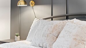 A neatly made bed with white linens and patterned pillows, a bedside lamp, and a small potted plant on a nightstand.